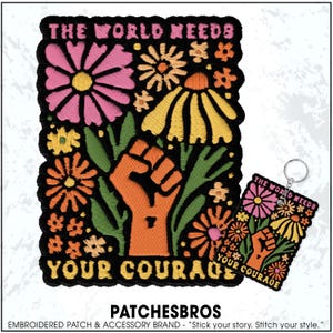 May include: Embroidered patch and keychain featuring a floral design with a raised fist. The patch and keychain have the text "THE WORLD NEEDS YOUR COURAGE" in a stylized font. The design includes pink, yellow, and orange flowers on a black background.