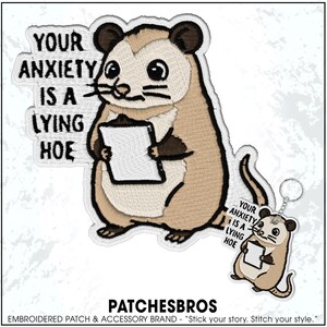 May include: Embroidered patch and keychain set featuring a cartoon opossum holding a clipboard. The patch and keychain have the text "YOUR ANXIETY IS A LYING HOE". The opossum is beige, brown, and white with black accents.