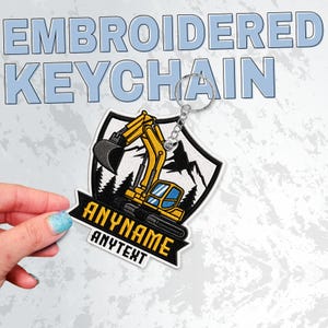 May include: A shield-shaped embroidered keyring featuring a yellow excavator, black pine trees, and mountain silhouettes. A black banner across the bottom displays the text ANYNAME and ANYTEXT in yellow capital letters with a metal ring attached.