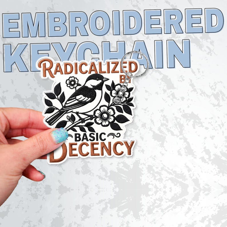 May include: An embroidered keychain featuring a black and white bird perched on a branch with flowers. The text reads RADICALIZED BY BASIC DECENCY in brown lettering. The keychain includes a silver metal ring attached to the top of the design.
