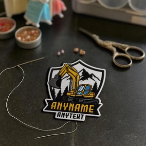 May include: A shield-shaped embroidered patch featuring a yellow excavator, black mountain silhouettes, and evergreen trees. The patch includes the text ANYNAME in yellow and ANYTEXT in white on a black background, with a needle and thread nearby.