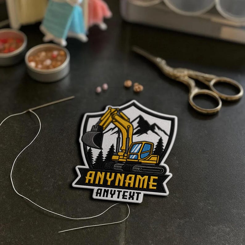 May include: A shield-shaped embroidered patch featuring a yellow excavator, black mountain silhouettes, and evergreen trees. The patch includes the text ANYNAME in yellow and ANYTEXT in white on a black background, with a needle and thread nearby.