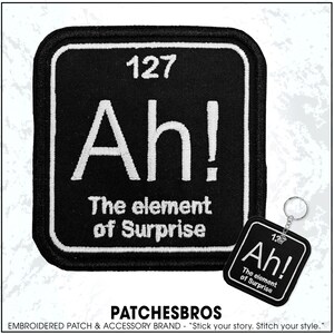 May include: Black square patch with white text reading "127 Ah! The element of Surprise." A matching keychain is also present. The patch and keychain have a white border.