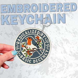 May include: A circular embroidered keychain featuring a robin perched on a branch with orange flowers. The design is set against a dark blue background with a white border. The text RADICALIZED BY BASIC DECENCY is stitched in white around the edge.