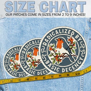 May include: Three circular embroidered patches featuring a robin perched on a branch with orange berries. The text RADICALIZED BY BASIC DECENCY circles the bird. A yellow measuring tape shows sizes ranging from 2 to 9 inches on a denim background.