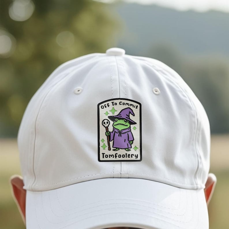 May include: A white baseball cap featuring a patch with a cartoon frog dressed as a wizard, holding a staff. The patch reads "Off To Commit Tomfoolery". The frog is green and wears a purple robe and hat.