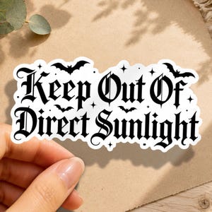 May include: A white die-cut sticker featuring the black text Keep Out Of Direct Sunlight in a gothic font. The design is decorated with small black bat silhouettes and four-pointed star icons scattered around the lettering on a white background.
