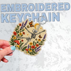 May include: An embroidered keyring featuring a skeleton surrounded by colourful flowers, mushrooms, and butterflies on a beige background. The design includes a yellow crescent moon and is attached to a silver metal ring. The text EMBROIDERED KEYCHAIN is visible in the background.