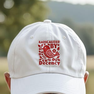 May include: A white baseball cap with a red graphic of a bird and the words "RADICALIZED BY Basic Decency". The cap has a curved brim and a button on top. The background is blurred.