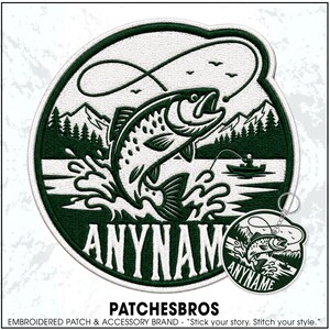 May include: A circular embroidered patch and a matching circular keychain featuring a dark green and white design of a fish jumping out of water, a fishing rod, a small boat with a person, and mountains. The text ANYNAME is printed below.