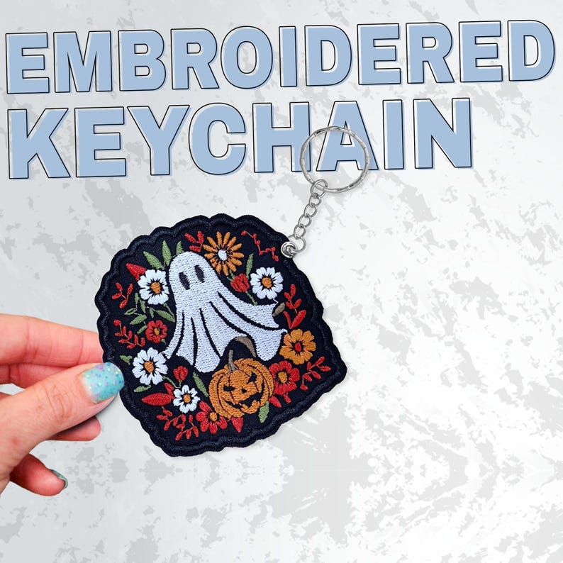 May include: Embroidered keychain featuring a ghost, pumpkin, and floral design. The keychain has a black background with white, orange, and red accents. The text "EMBROIDERED KEYCHAIN" is displayed in light blue.