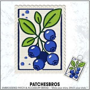 May include: A rectangular embroidered patch featuring a cluster of blue blueberries with green leaves on a cream background, framed by a dark blue border and a scalloped edge. A smaller version of the same design is attached to a metal keyring.