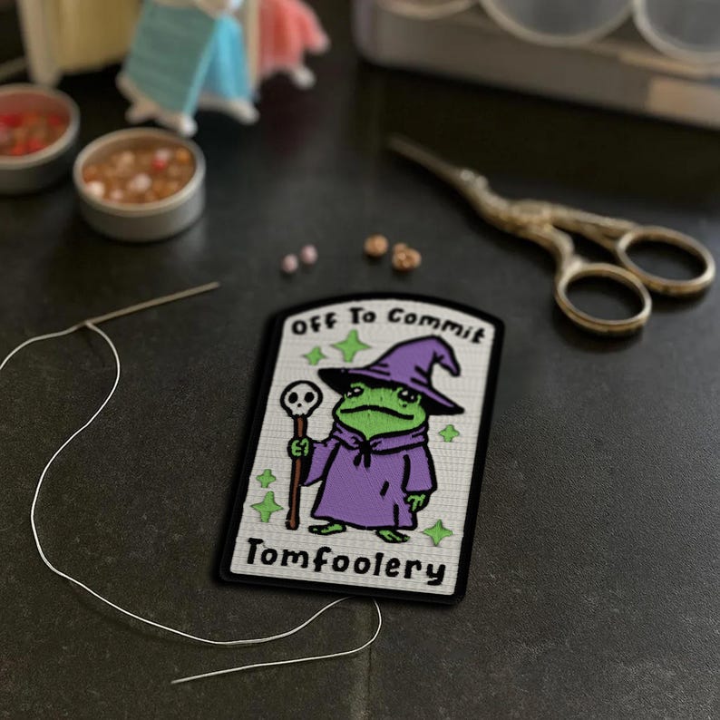May include: A rectangular patch featuring a cartoon frog in a purple wizard robe and hat, holding a staff with a skull. The text "OFF To Commit" and "Tomfoolery" are above and below the frog. Green stars and a needle with white thread are also visible.