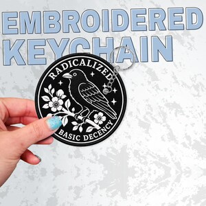 May include: A round black embroidered keychain featuring a white graphic of a bird perched on a flowering branch. The text RADICALIZED BASIC DECENCY is stitched in white along the circular border, accompanied by small star icons and a metal ring.