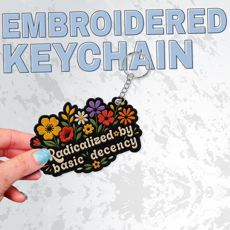 May include: A black embroidered keychain featuring a colorful floral arrangement with yellow, red, orange, and purple flowers. The text "Radicalized by basic decency" is stitched in a cream-colored font across the center of the floral design.