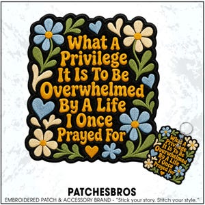 May include: A black embroidered patch and a matching keychain featuring the yellow text What A Privilege It Is To Be Overwhelmed By A Life I Once Prayed For, surrounded by light blue and cream flowers, green leaves, and small blue and yellow hearts.