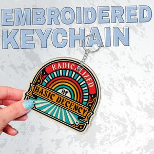 May include: Embroidered keychain with a colorful patch. The patch features a rainbow design with the words "RADICALIZED" and "BASIC DECENCY". The keychain is attached to a silver metal ring.