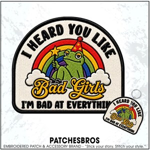May include: An embroidered patch and a matching keychain featuring a green frog wearing a party hat and holding a yellow star wand. The design includes a rainbow, white clouds, and the text I HEARD YOU LIKE Bad Girls I'M BAD AT EVERYTHING.