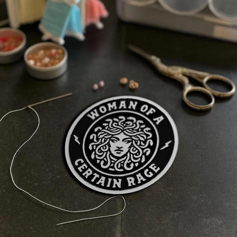 May include: A black and white patch with the text "WOMAN OF A CERTAIN RAGE" encircling a Medusa illustration. A needle and thread, small containers, and scissors are also visible.