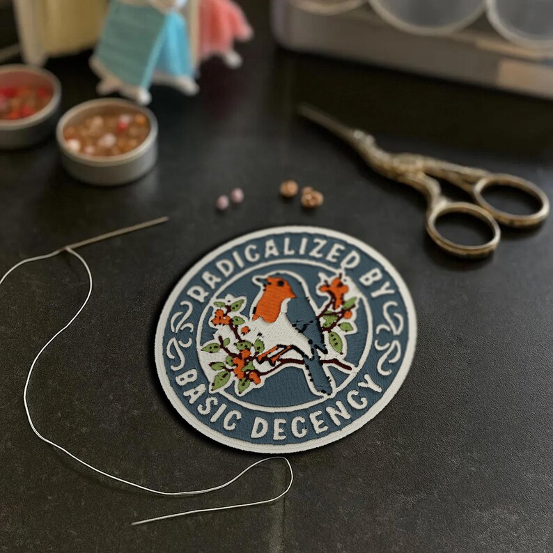 May include: A circular embroidered patch featuring a blue robin perched on a branch with orange berries. The design is surrounded by a white border with the text RADICALIZED BY BASIC DECENCY in dark blue lettering on a light blue background.