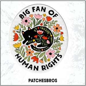 May include: Embroidered patch with a black cat curled up in a floral design. The patch is oval-shaped and features the text "BIG FAN OF HUMAN RIGHTS". The design includes pink, orange, and yellow flowers with green leaves. The brand name "PATCHESBROS" is at the bottom.