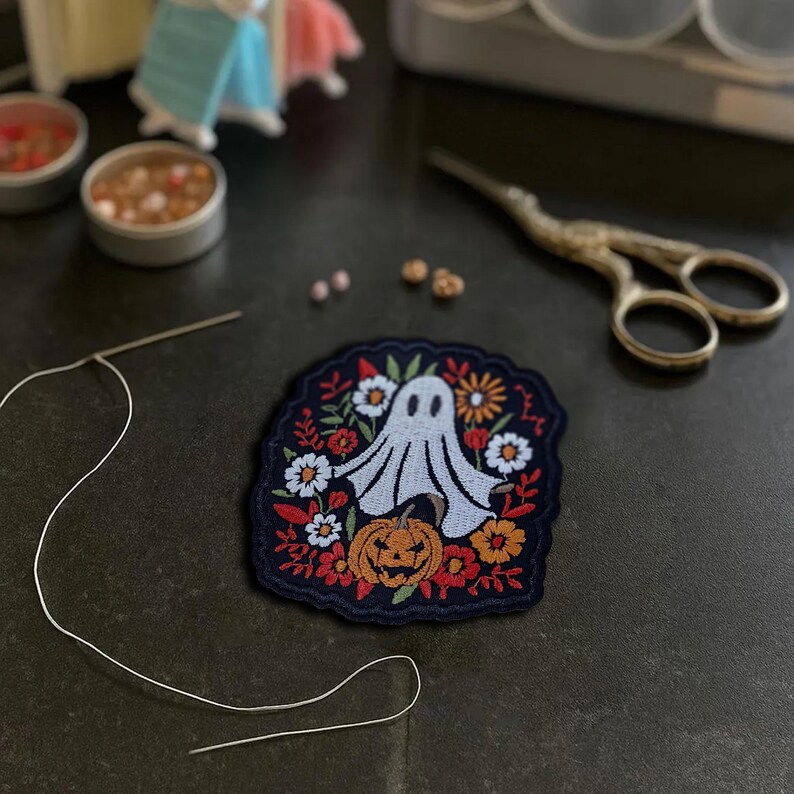 May include: Embroidered patch featuring a friendly ghost, pumpkin, and floral elements in orange, white, and red on a navy blue background. The patch is surrounded by sewing tools, including scissors and a needle with thread, suggesting a craft project.