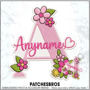 May include: An embroidered patch shaped like the letter A in light pink with white accents, decorated with pink flowers and green leaves. The word Anyname is stitched in dark pink cursive across the center, ending with a small heart shape.