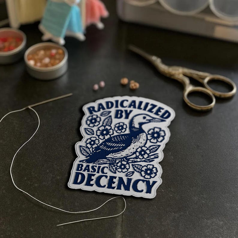 May include: A white patch with a navy blue loon illustration and the text "RADICALIZED BY BASIC DECENCY". The patch is surrounded by white flowers. A needle and thread, scissors, and small containers with beads are also visible.