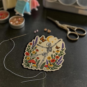 May include: An embroidered patch featuring a white skeleton dancing amidst colourful wildflowers, red and white mushrooms, orange butterflies, and a yellow crescent moon on a cream-coloured fabric background with a scalloped edge.