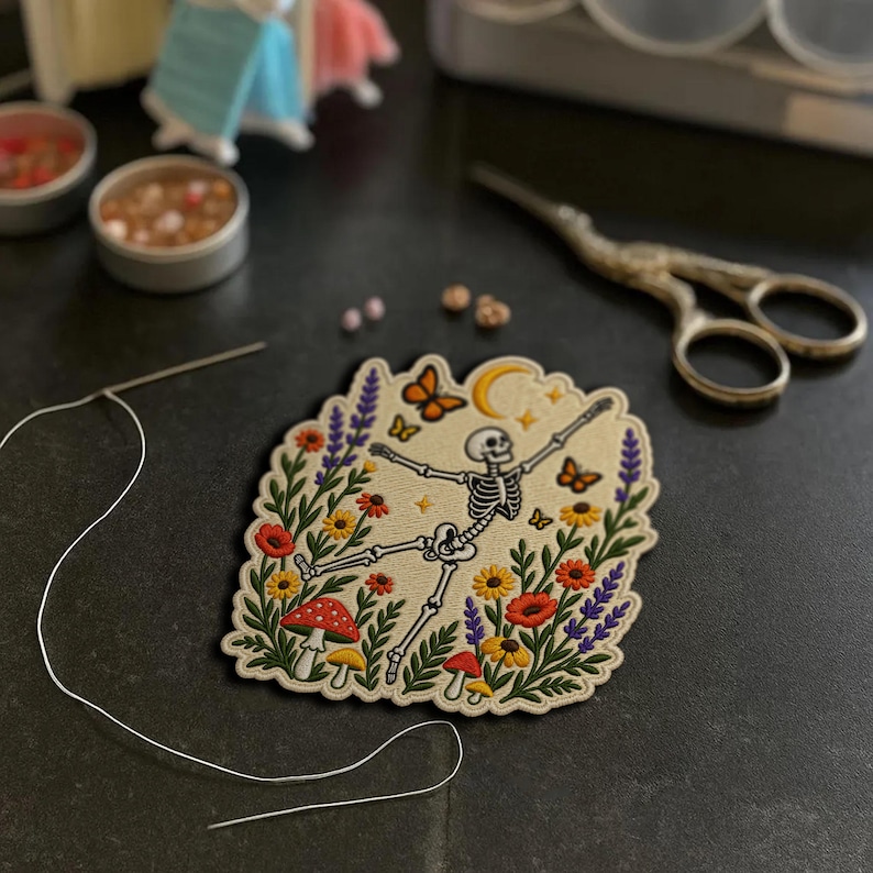 May include: An embroidered patch featuring a white skeleton dancing amidst colourful wildflowers, red and white mushrooms, orange butterflies, and a yellow crescent moon on a cream-coloured fabric background with a scalloped edge.