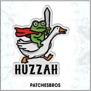 May include: Embroidered patch featuring a green frog wearing a red cape and holding a sword, riding a white goose. The word "Huzzah" is embroidered below the image, with "PATCHESBROS" below that.