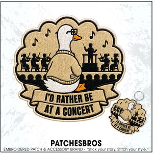 May include: An embroidered patch and matching keychain featuring a white duck wearing glasses and a tan sweater, sitting before an orchestra with a conductor. The design includes black musical notes and a banner with the text I'D RATHER BE AT A CONCERT.