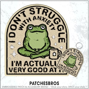 May include: Embroidered patch with a green frog design, text reads "I don't struggle with anxiety, I'm actually very good at it." A matching keychain is also shown. The patch is beige with black and green stitching.