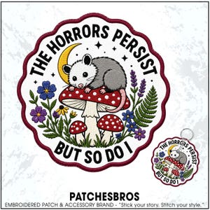 May include: A circular embroidered patch featuring a gray opossum resting on a red and white spotted mushroom under a yellow crescent moon. The text THE HORRORS PERSIST BUT SO DO I surrounds the design, which is accented by blue and purple flowers.