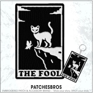 May include: Black and white embroidered patch and keychain featuring a cat standing on a cliff with a butterfly, labeled "THE FOOL." The patch and keychain are part of a tarot card-themed accessory collection.