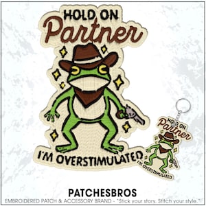 May include: Embroidered patch and keychain set featuring a green frog wearing a cowboy hat and holding a gun. The patch and keychain read "HOLD ON Partner" and "I'M OVERSTIMULATED". The patch has a cream-colored border with gold accents.