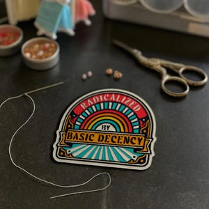 May include: A colorful patch with the text "RADICALIZED BY BASIC DECENCY" is the focal point. The patch features a rainbow design and is surrounded by sewing supplies, including a needle, thread, and small scissors. The patch is on a dark surface.