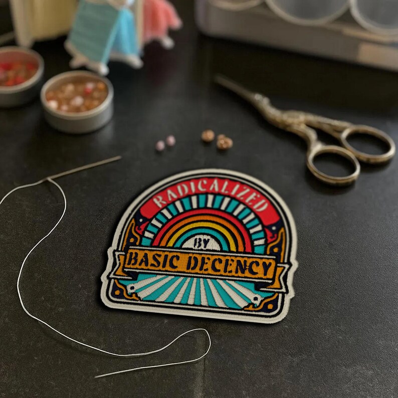 May include: A colorful patch with the text "RADICALIZED BY BASIC DECENCY" is the focal point. The patch features a rainbow design and is surrounded by sewing supplies, including a needle, thread, and small scissors. The patch is on a dark surface.