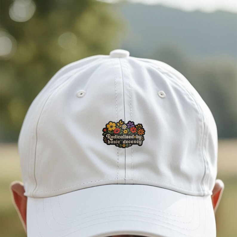 May include: A white baseball cap featuring a central embroidered patch with a colorful floral design and the text "Radicalized by basic decency" in a dark, stylized font. The cap has a structured crown, a curved brim, and ventilation eyelets.