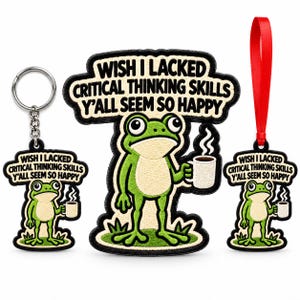 May include: A keychain, a patch, and an ornament featuring an embroidered green frog holding a white coffee mug. The text reads: WISH I LACKED CRITICAL THINKING SKILLS Y'ALL SEEM SO HAPPY. The keychain has a metal ring, and the ornament has a red ribbon.