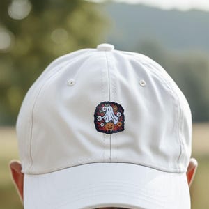 May include: A white baseball cap with a Halloween-themed patch. The patch features a ghost, a jack-o'-lantern, and colorful flowers. The cap is made of a smooth material and has a curved brim.