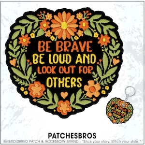 May include: Embroidered patch with the text "BE BRAVE BE LOUD AND LOOK OUT FOR OTHERS" in orange and yellow. The patch is black with a floral wreath design in orange and green. A matching keychain is also visible.