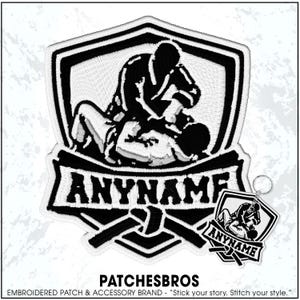 May include: A black and white embroidered patch and matching keychain featuring a shield design with two figures grappling in martial arts uniforms. The text ANYNAME is embroidered in a bold font across the center banner above a tied belt.