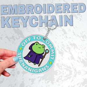 K&ouml;nnte beinhalten: Gestickter Schl&uuml;sselanh&auml;nger mit einem Cartoon-Frosch in einem lila Gewand, der einen Stab h&auml;lt. Der runde Schl&uuml;sselanh&auml;nger tr&auml;gt den Text "OFF TO COMMIT SHENANIGANS". Er hat einen silbernen Schl&uuml;sselring.