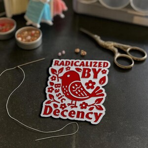 May include: A red and white patch with the text "RADICALIZED BY Basic Decency" and a bird illustration. The patch is surrounded by floral designs. A needle and thread, small tins, and scissors are also visible.