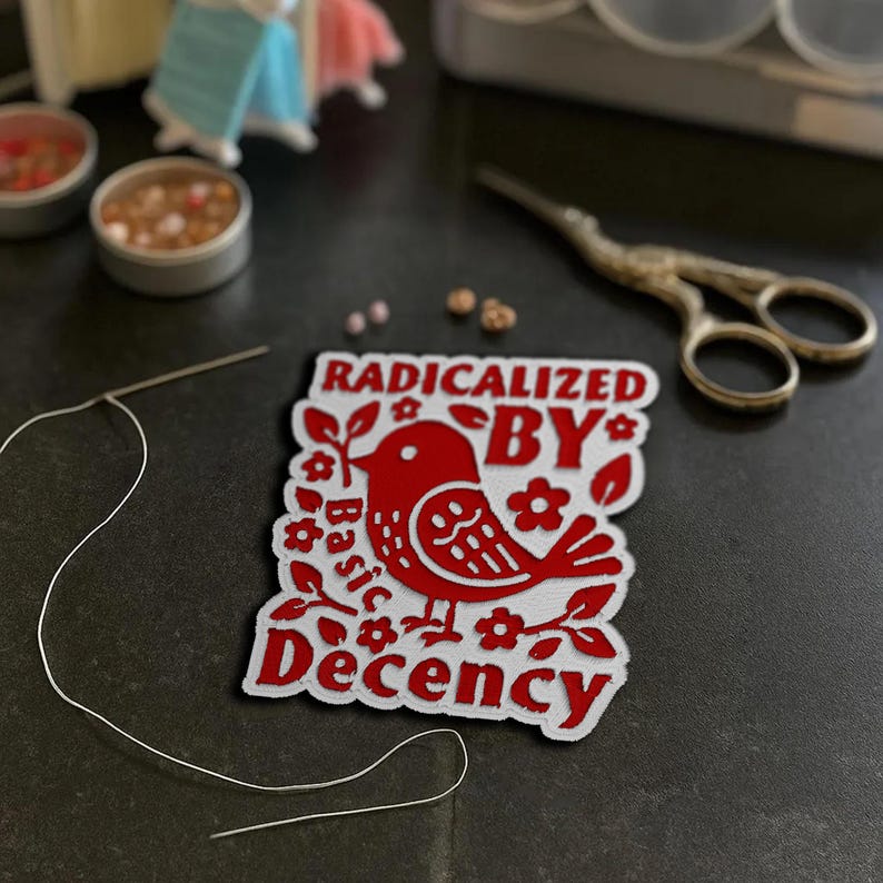 May include: A red and white patch with the text "RADICALIZED BY Basic Decency" and a bird illustration. The patch is surrounded by floral designs. A needle and thread, small tins, and scissors are also visible.