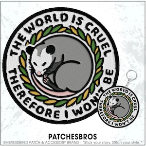 May include: Embroidered patch featuring a curled opossum within a laurel wreath. The text "THE WORLD IS CRUEL THEREFORE I WON'T BE" encircles the design. A matching keychain is also visible.
