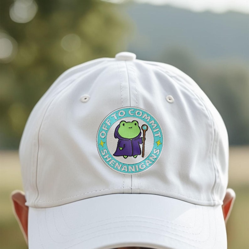 K&ouml;nnte beinhalten: Eine wei&szlig;e Baseballkappe mit einem runden Aufn&auml;her mit einer Cartoon-Fr&ouml;schin in einem lila Gewand, die einen Stab h&auml;lt. Der Aufn&auml;her tr&auml;gt die Aufschrift "OFF TO COMMIT SHENANIGANS" in einem t&uuml;rkis-wei&szlig;en Design.