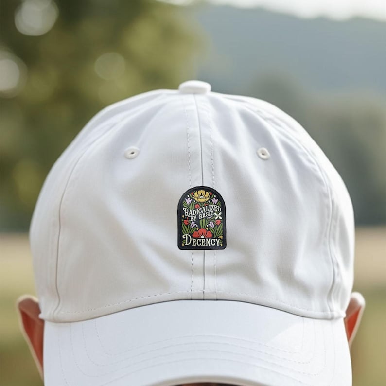 Pu&ograve; includere: Un cappellino da baseball bianco con visiera curva. Il cappellino presenta una toppa nera con le parole "RADICALIZED BY BASIC DECENCY" e illustrazioni floreali in rosso, giallo e verde.