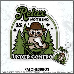 May include: Embroidered patch and keychain set featuring a raccoon in sunglasses and a hat, relaxing in a camping chair. The patch and keychain have the text "Relax, Nothing Is Under Control" in a green and white design.